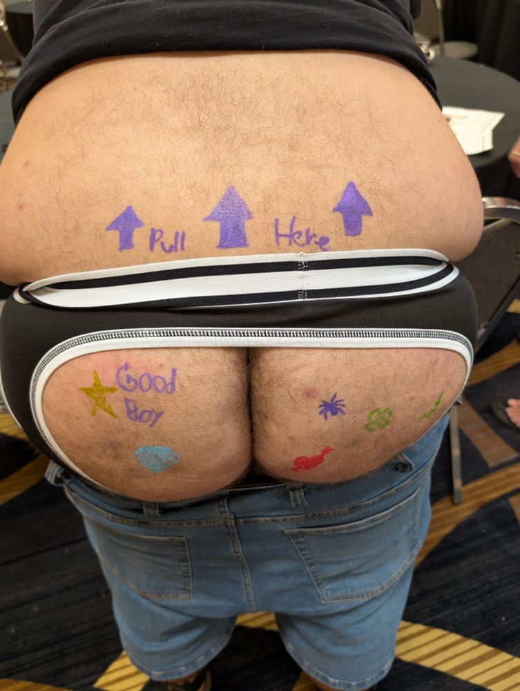 Close up rear view of me wearing a black t-shirt, blue jean shorts, and assless briefs with several doodles on my butt, including the words "Good Boy" and, on my lower back, "Pull Here" with three arrows pointing up