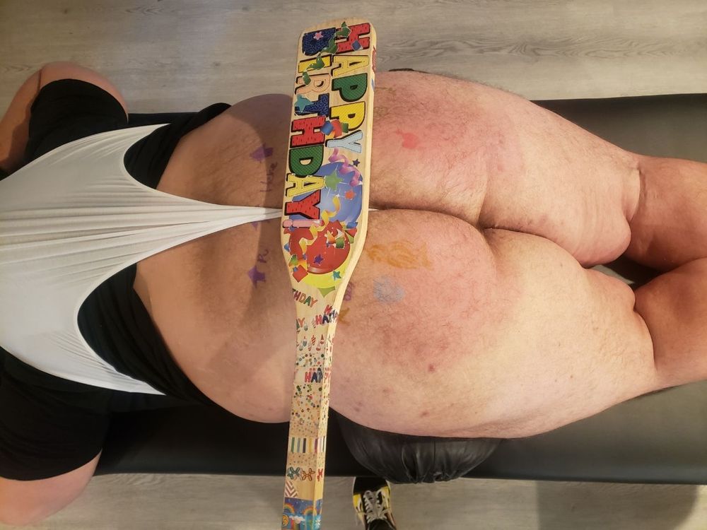 "Ceiling cam" shot of me lying on my stomach on a massage table wearing a black t-shirt and white briefs that have been secured in an atomic wedgie, exposing my bare butt with smudged doodles and writing on it and SleepyBap's "Oarson Welts" paddle decorated with confetti and "Happy Birthday" stickers laying across my upper butt
