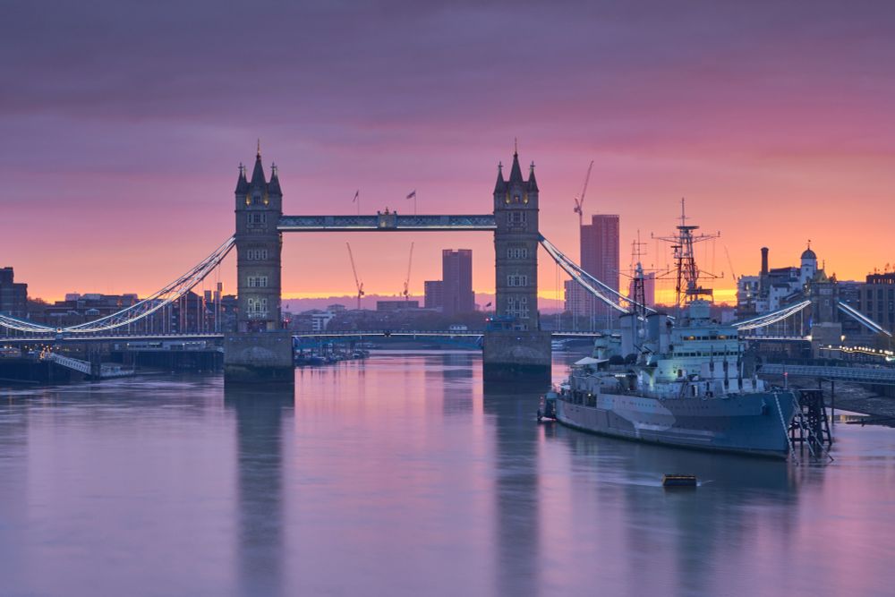 Tower Bridge and HMS Belfast at sunrise, the yellow glow on the horizon lighting the think clouds above to reveal oranges, pinks and purples 