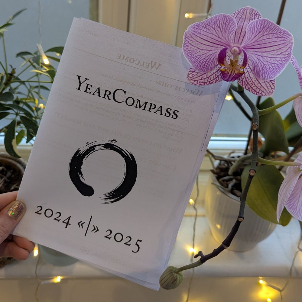 Photo of the Year Compass booklet for 2024/25 printed out, next to a pink flowering orchid and some fairy lights on a window sill