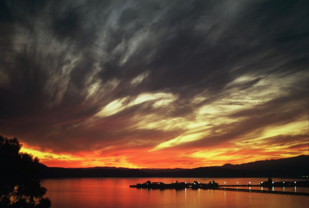 A fiery sunset over San Pablo Bay looking at the distant hills of Marin County, CA. Sky looks streaked with grey and white that morphs into fiery gold and orange that reflects in the water. In the foreground, the Standard Oil longwharf looks pretty good for a change.