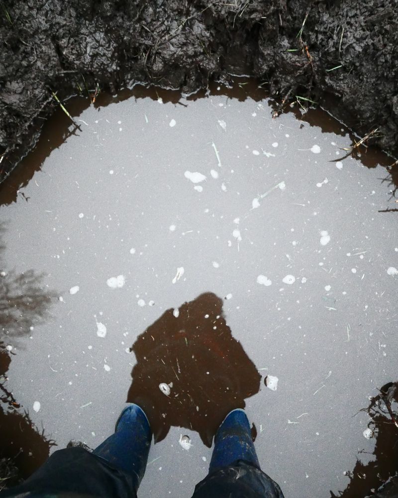 Je kijkt van bovenaf naar twee benen en voeten met blauwe laarzen die in een ronde poel staan. de persoon wordt in het water weerspiegeld.