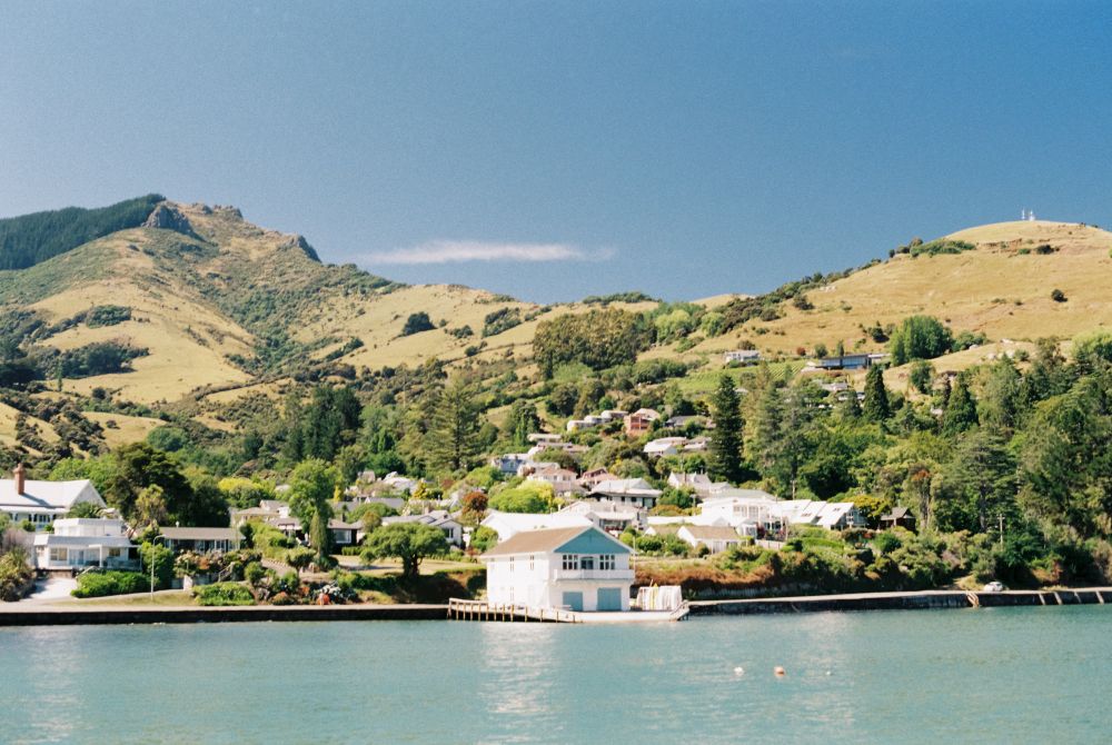 A photo of a hills wth two distinct peaks, partially covered in trees, bushes and some houses. A white building with blue doors is situated right in the centre at the base of the hills on the edge of a body of water.
