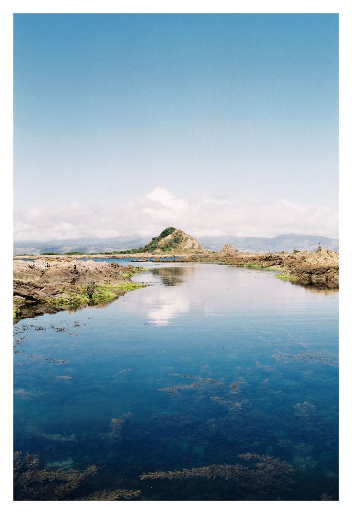 A photo of a rocky coastal area with a shallow pools of water forming a mini river, filling the lower central half of the frame. The water is calm, cleanly reflecting the blue skies and small island in the upper half of the frame. The rocks on the sides of the water pool are greenish, being covered in moss. Near the camera where the reflections fade out, underwater plants can be seen through the water.