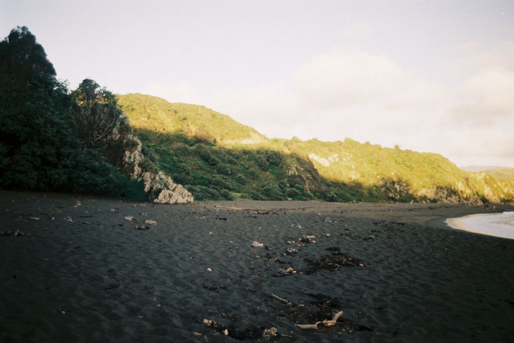 A photo of Breaker Bay, standing on the beach. It looks towards green hills bordering the beach that are lit at the tops by golden light from a setting sun.