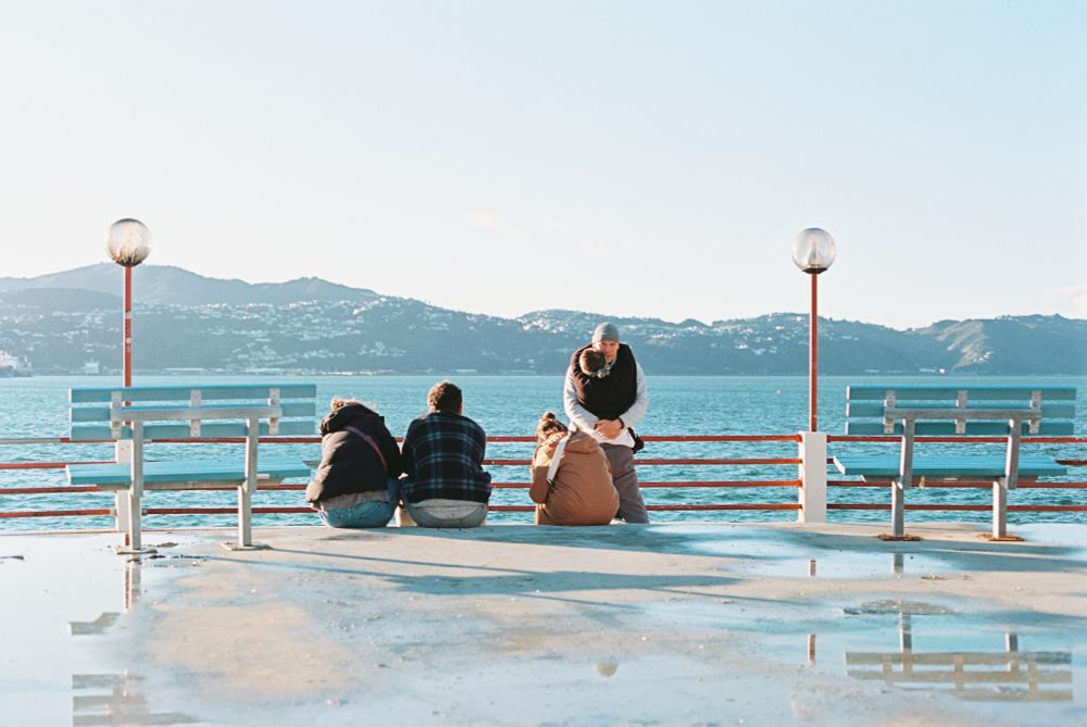 A photo of a group of people on rooftop balcony near sunset. Two people are hugging. The floor is concrete with small puddles reflecting the sky and benches on the balcony. The sky is clear with no clouds. The photo is slightly overexposed, giving it a dreamlike quality.