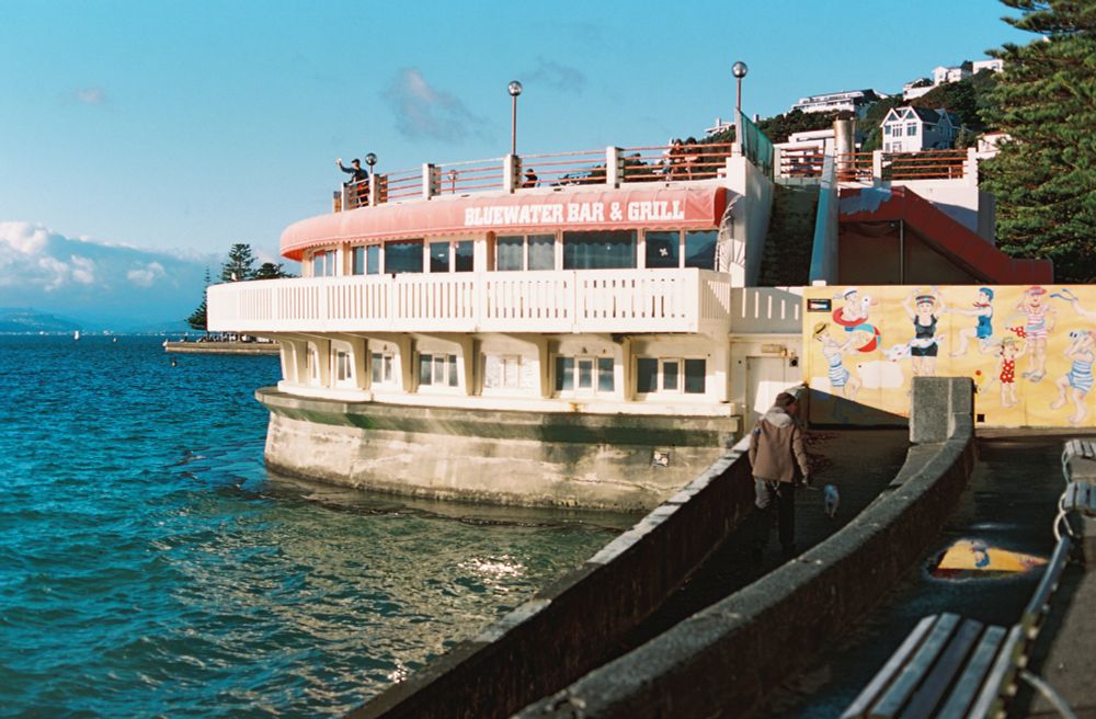 A photo of a 1980s style rotunda on the edge of Oriental Bay near sunset. The rotunda has windows in the bottom section, a fenced balcony in the middle and a rooftop balcony at the top. A red awning near the top has white text reading “Bluewater Bar & Grill”.