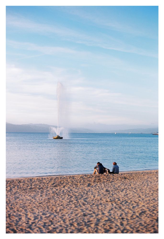 A photo of a beach overlooking Oriental Bay lit by reddish light from a setting sun. Off the coast is a large geyser-like fountain. Two people are sitting at the edge of the water watching it.