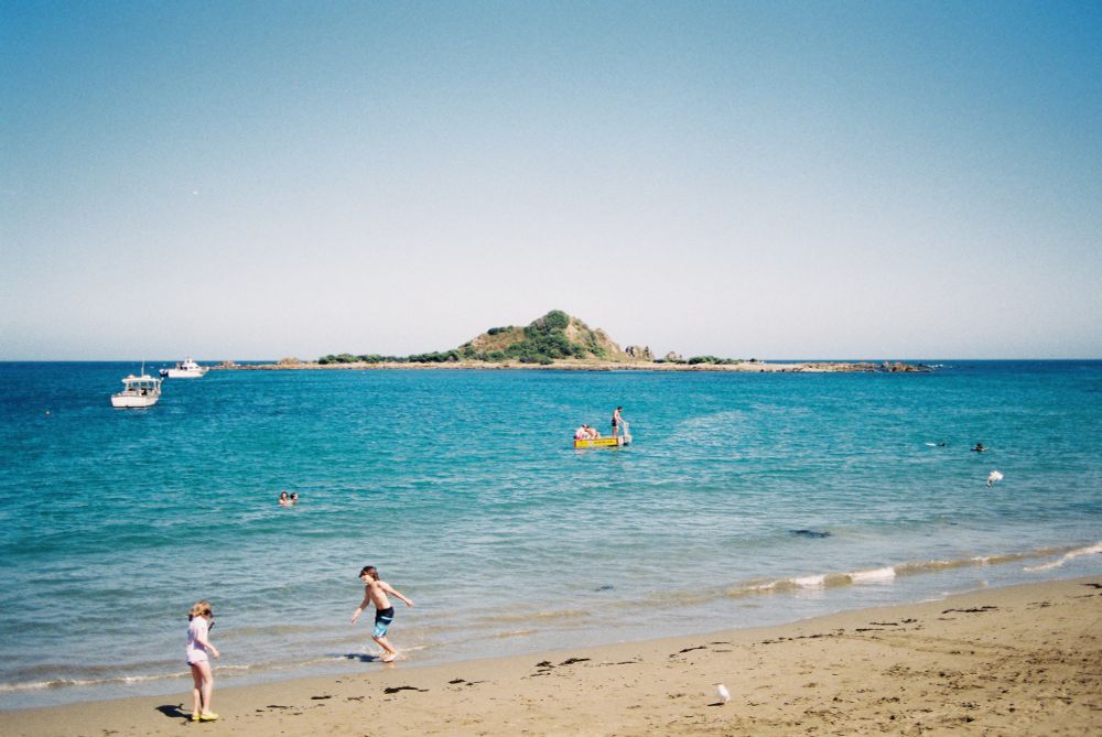 A photo of a beach on a bright sunny day with clear skies. A symmetrical island can be seen centred in frame in the distance. The water is a rich teal with small yellow and white boats floating on it. In the foreground on the sand, two children are playing.