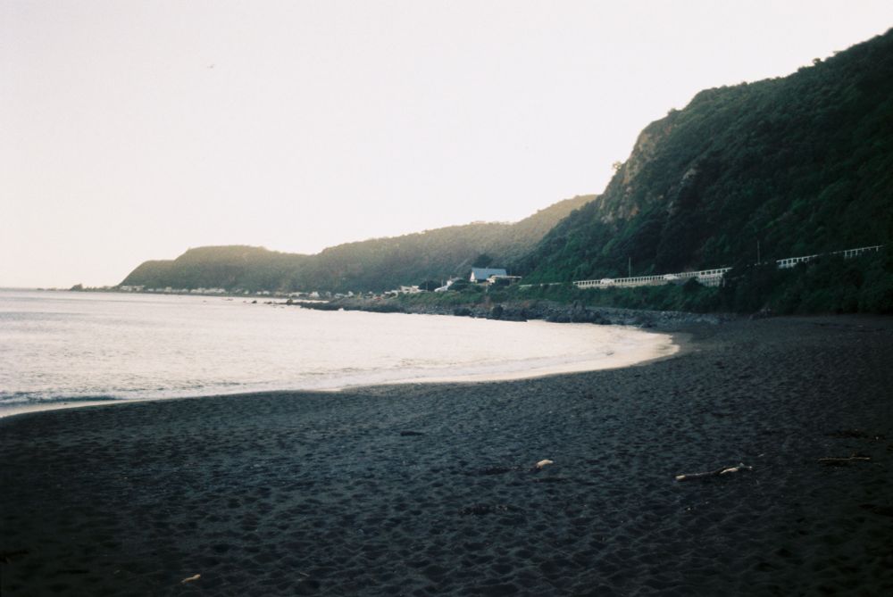 A photo of Breaker Bay on a beach with dark-grey sand. The shot looks towards some steep hills as the sun sets behind them, creating soft crepuscular rays.