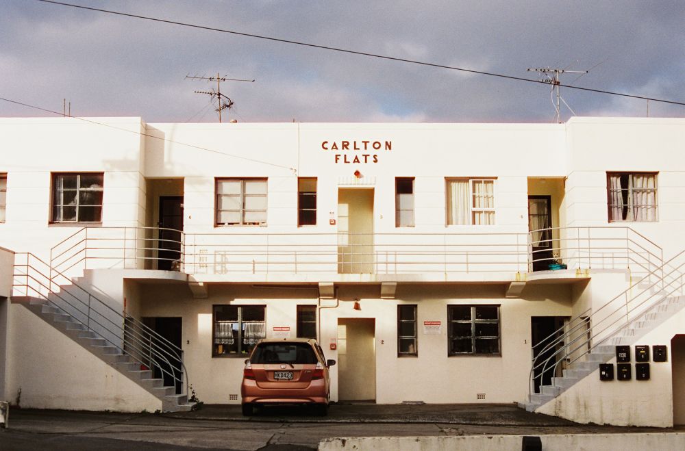 A photo of a symmetrical white building centred in frame. The building has text at the top centre reading ‘Carlton Flats’. There are two wings on either side with staircases leading up to a balcony in the middle.
