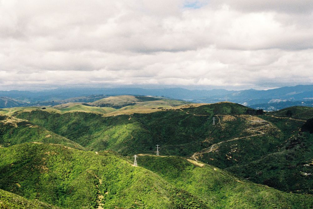 A photo of a series of green hills covered in bush and trees. The hills are partially lit by light streaming through broken clouds.