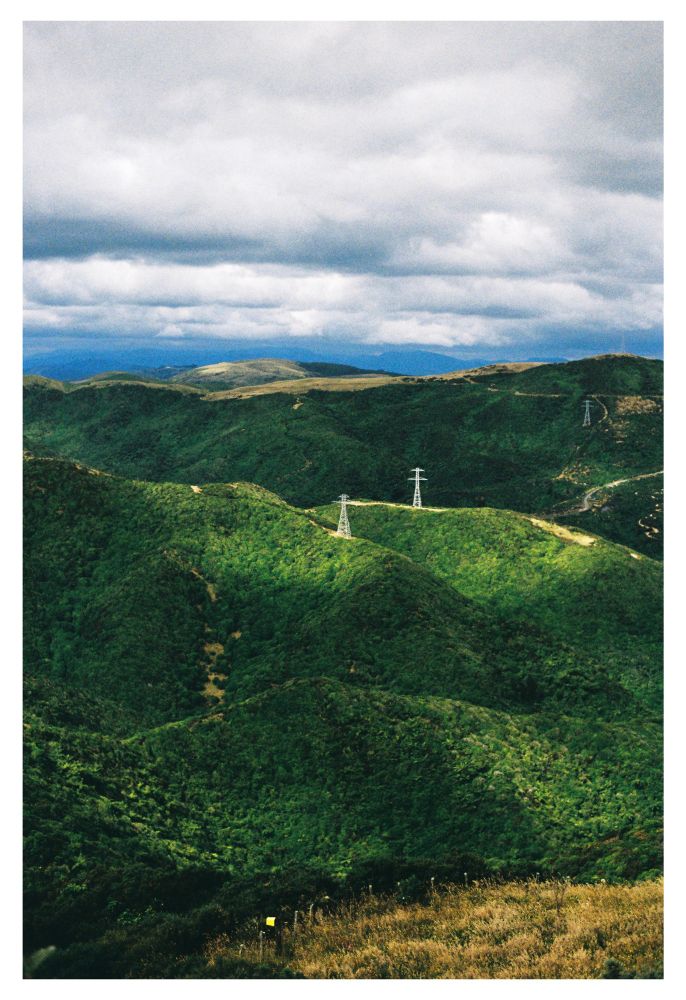 A photo of a series of hills from a distance at a high vantage point. The hills are dark green and covered in dense trees and bush. Two power pylons can be seen on the central two peaks, lit by light peaking through the clouds.