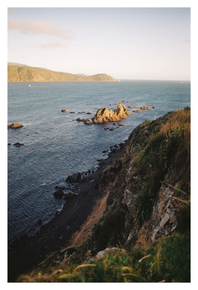 A photo taken from the edge of a cliff, overlooking a large bay, with a a headland in the far distance. A group of sharp rocks stick out of the water in the middle of the frame, lit by light from a setting sun.