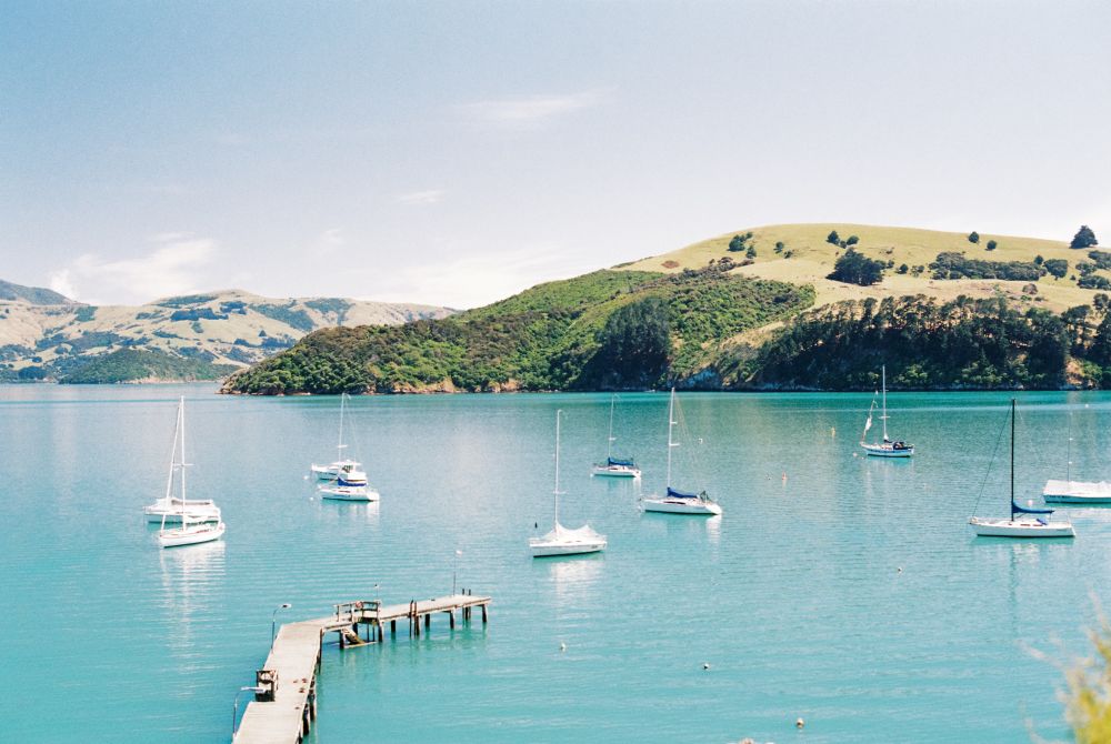 A photo overlooking a body of water with ten boats scattered around the frame. A small dock is partially visible  coming from the lower left of the frame. Tree covered hills can be seen in the distance.