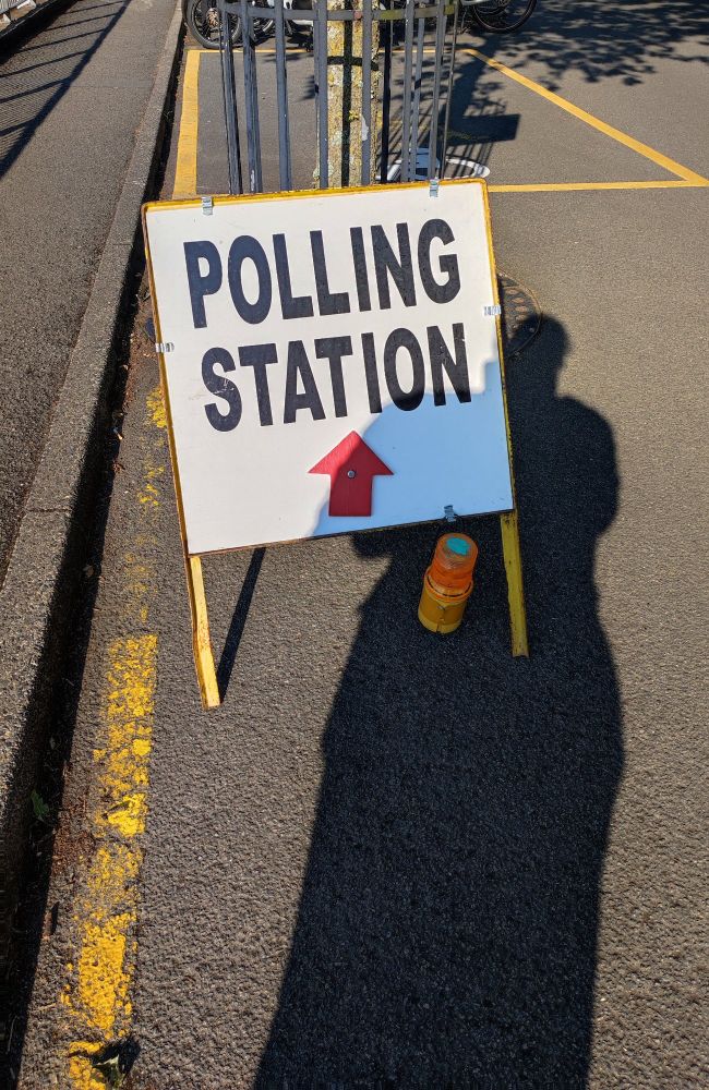 A photo of a roadside sign reading "POLLING STATION". There is a shadow cast over it by the photographer.