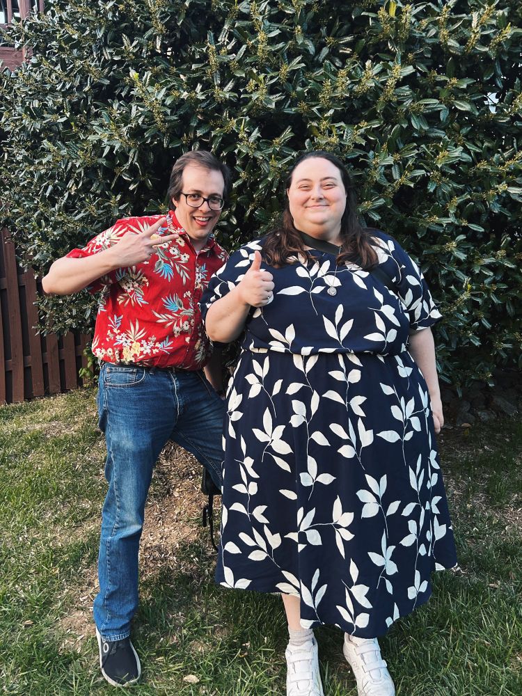 Kate and her friend Dan stand in front of a giant green bush. Dan is holding up a peace sign and Kate is doing a thumbs up.