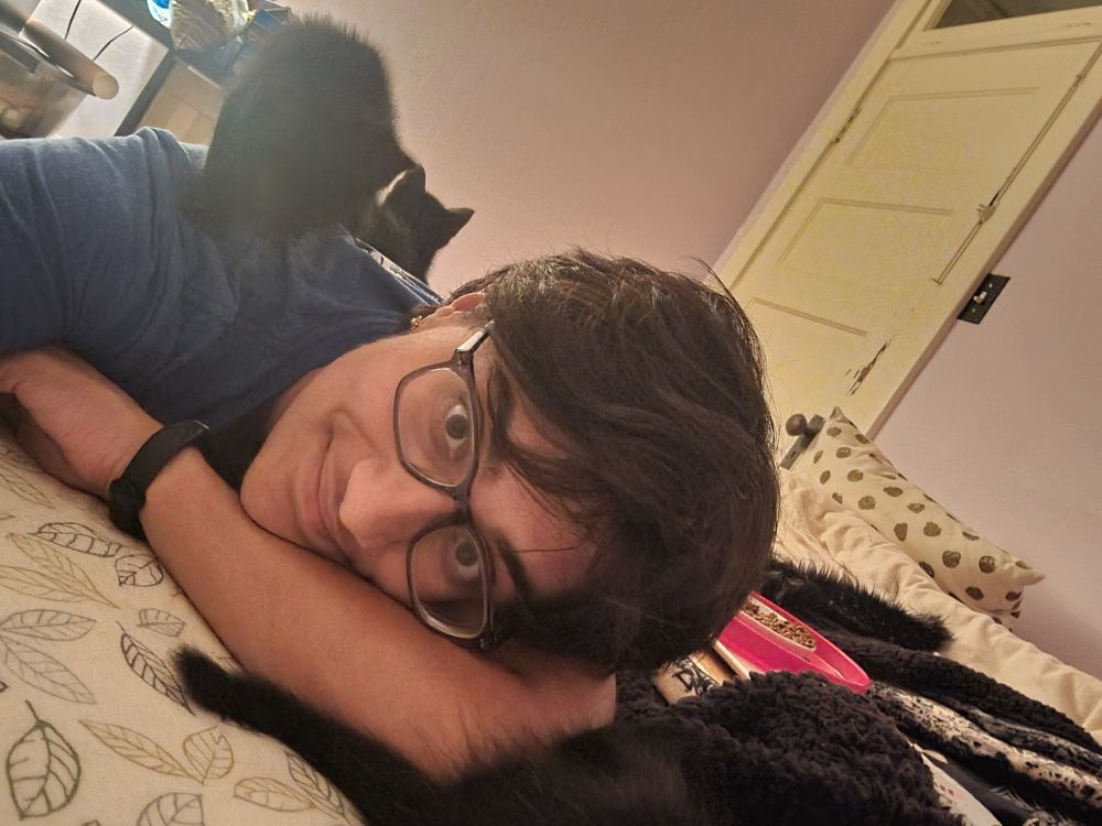 Me, lying on my stomach on a bed. On my back are 2 black kittens, both standing. On front of me, half out of frame, is a third black kitten. 