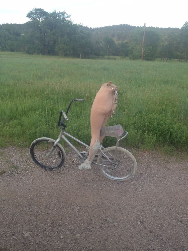 the bottom half of a mannequin, seated on an old bicycle, with jingle bells attached at the top sits on the side of a dirt road with grass and trees in the background
