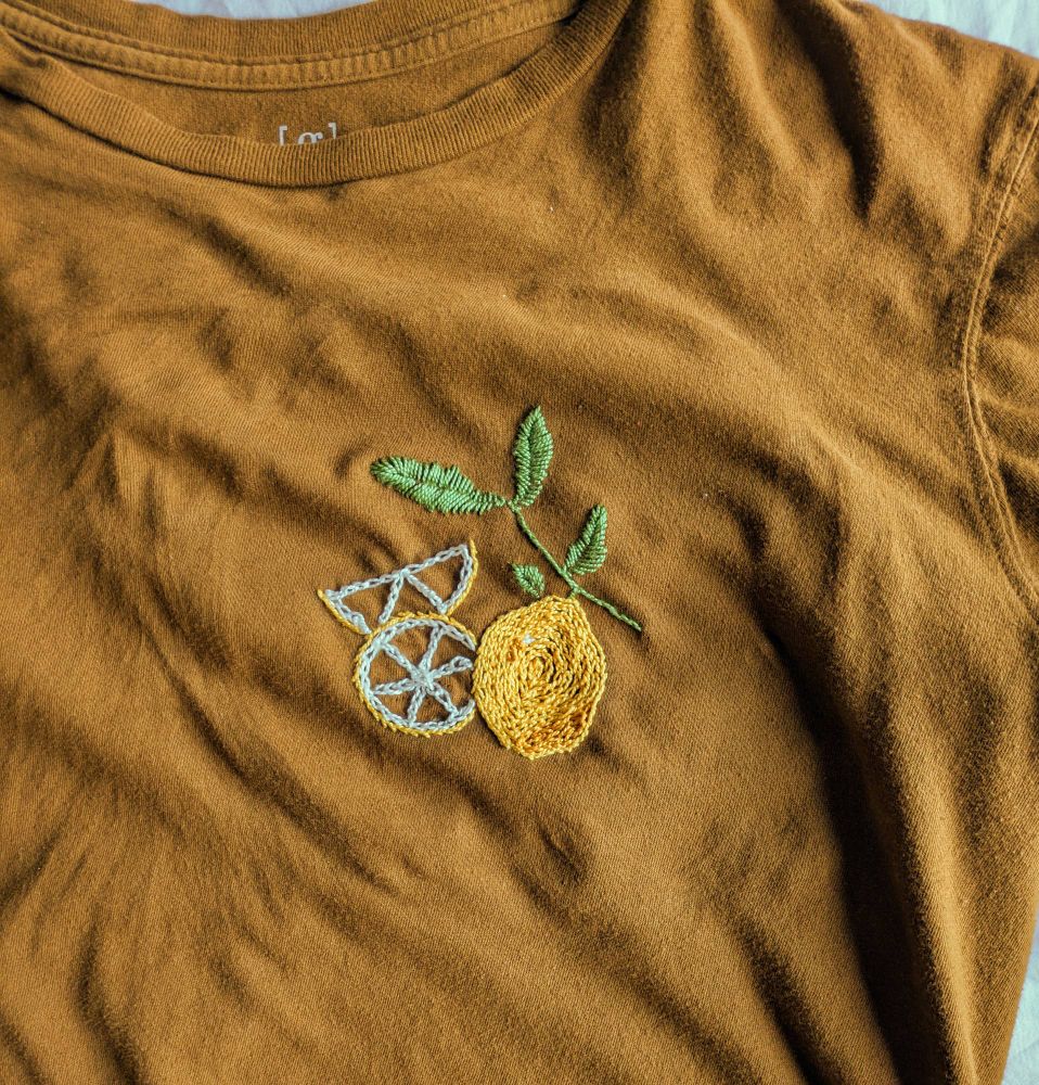 An embroidery design on a dark orange/brown shirt of a lemon and a branch with leaves. There is a lemon, a whole lemon slice, and a lemon wedge.