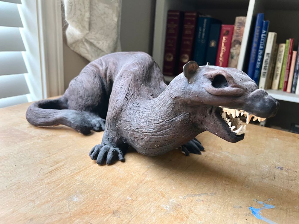 Sculpture of an angry weasel with no eye, on a wooden desk in front of a book shelf