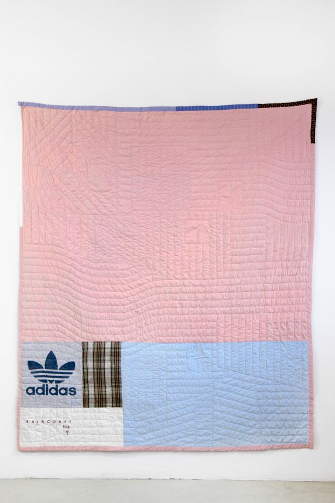 Pieced back of a quilt in pink, blue, brown plaid, and white, with one block made from an Adidas shirt in grey, hung on a white wall.