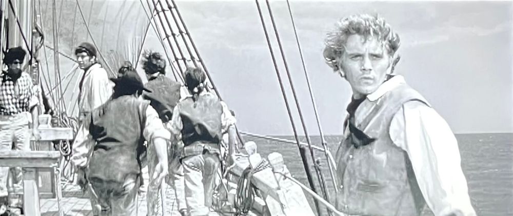 As sailors fall in to line the deck of the merchant vessel Rights of Man, the foretopman Billy Budd turns at the sound of his name. Occupying the right-hand third of the frame in a medium close-up is 23-year-old Terence Stamp in his first leading role.