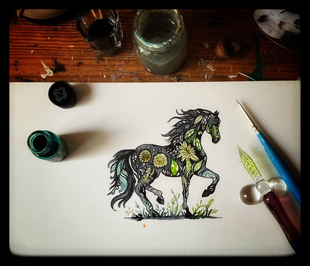 An ink doodle of a horse that has green leaves and squiggles in it. Nearby lies an ink pen and a paintbrush. To the left an open bottle of green ink. At the top a small jar of dirty water.