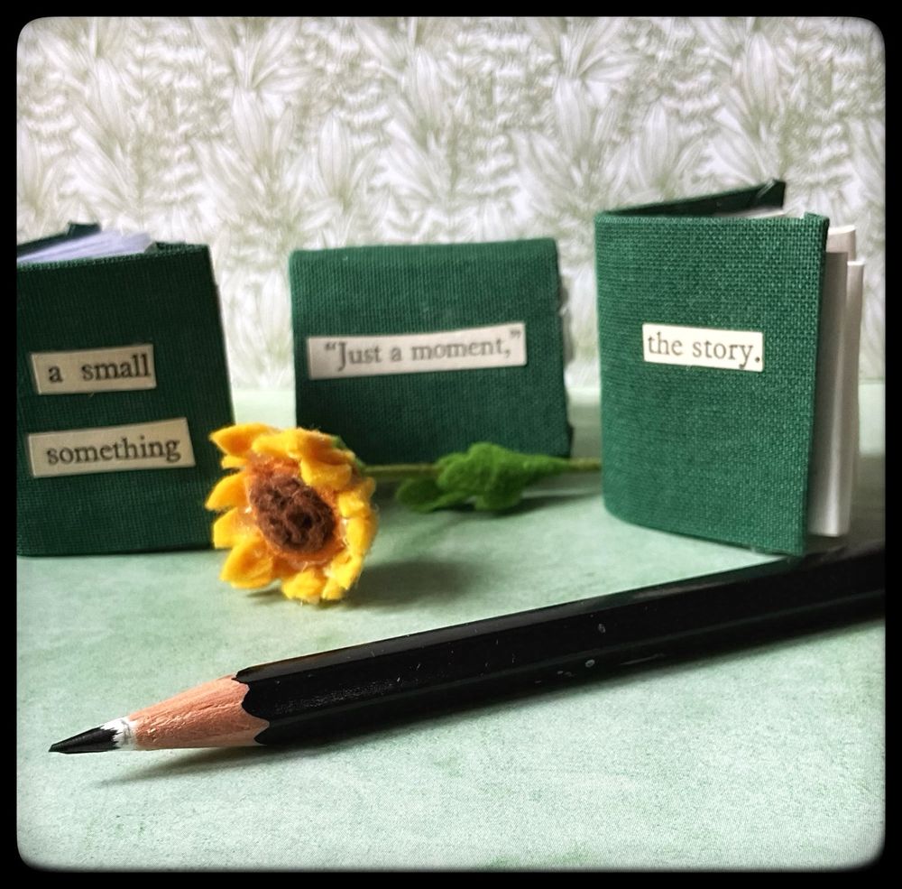 A black pencil lies in front of three green covered mini books. The left side book has the words “a small something” on it. The middle book has “just a moment” on it. The right hand side book has “ the story” on it. In the middle is a tiny felt sunflower.