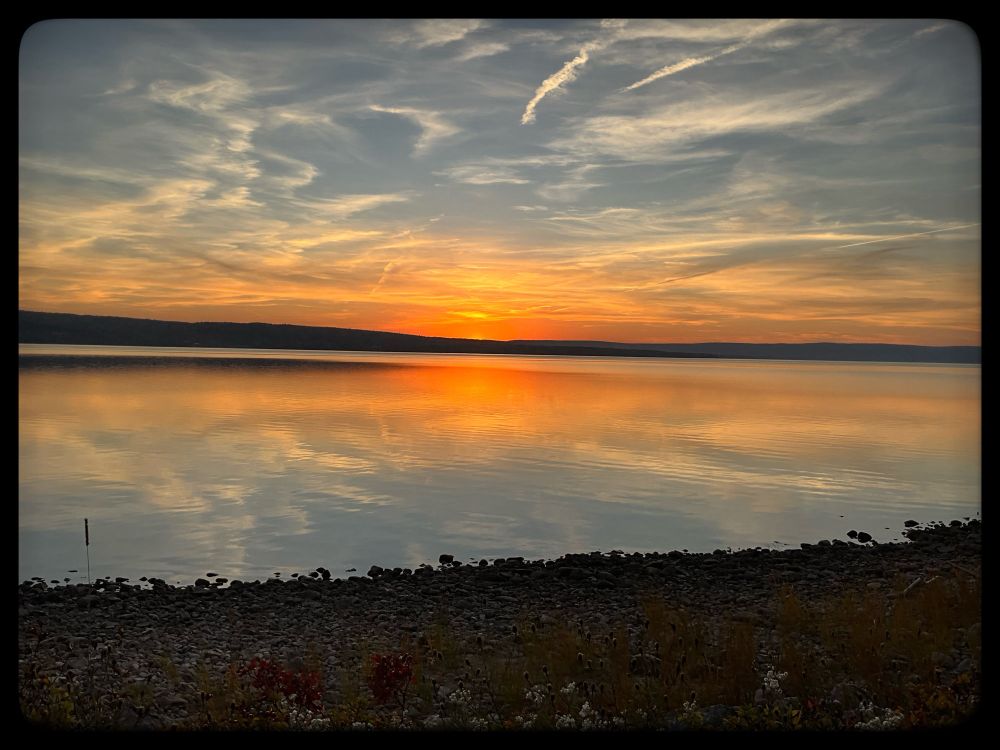 Orange streaks across blue grey are reflected in water. A thin slope of land divides the picture.