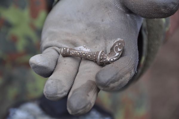 Photograph of a hand wearing a blue glove in the hand is a silver brooch about the width of the hand in size, it is silver and ornate.