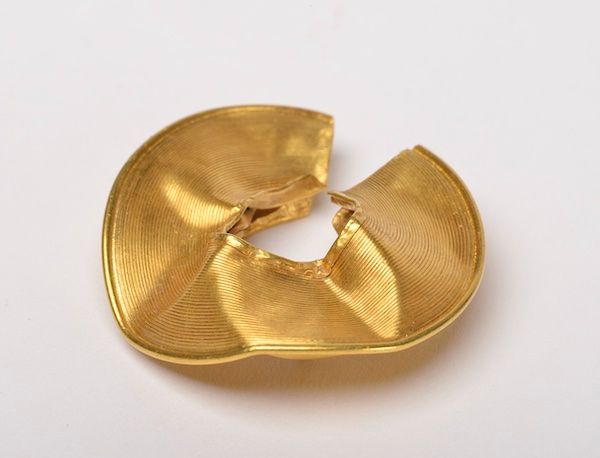 Photograph of a gold lock ring. Circular in shape it has a hole in the middle of the disc. It is damaged and dented.  
