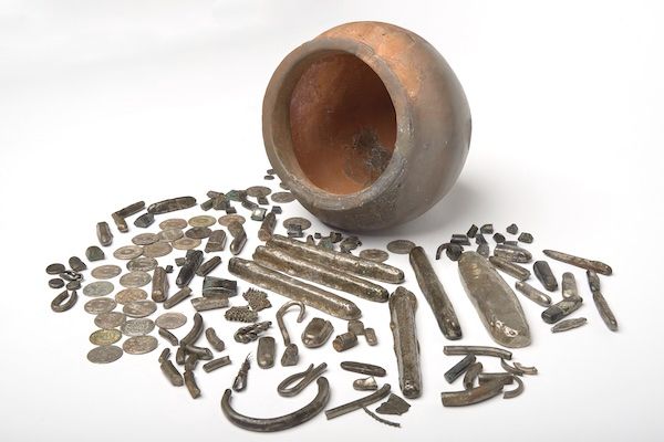 Photograph of an empty terracotta pot lying on its side. In front of the pot are an assortment of metal objects including coins, ingots and fragments of jewellery.