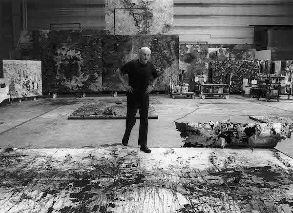 A black and white photograph of the artist Anselm Kiefer. It show a white male standing in the centre of the photograph in an artists studio. There are artworks on the floor and around the walls. He is standing with one foot on an artwork on the floor. He is wearing black trousers shoes and a black t-shirt. His hands are on his hips. 

The artist - Anselm Kiefer at work in his studio Photo courtesy Barbara Klemm