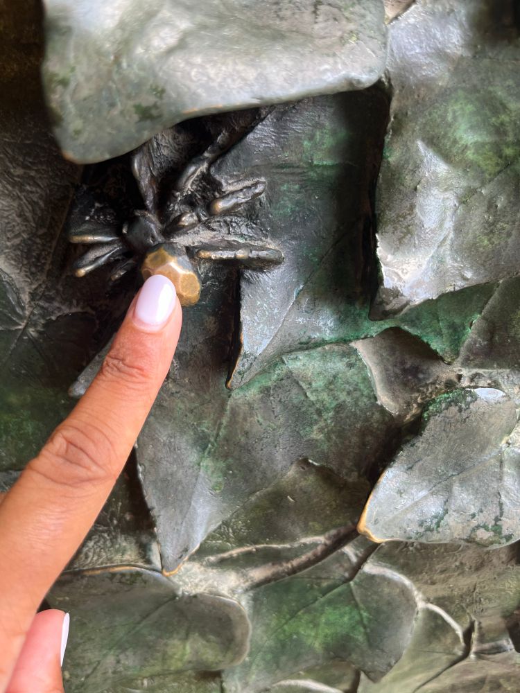 Metallic sculpted spider surrounded by green sculpted leaves. The surface of the spider’s abdomen is worn and shinier brass. A finger and thumb with brown skin and gray nail polish are gesturing to the spider.