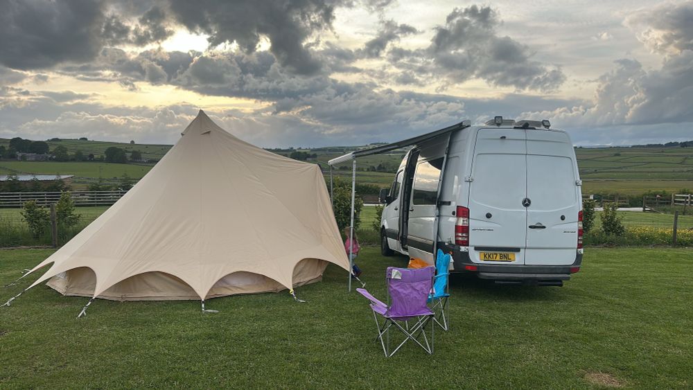 Bell tent, camper van and two camping chairs in a field in the Peak District. The sun is pushing through the clouds in the sky. 