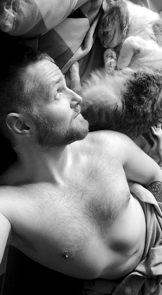 Shirtless bearded person cuddling on a couch with a cat. Photo is in high contrast black and white