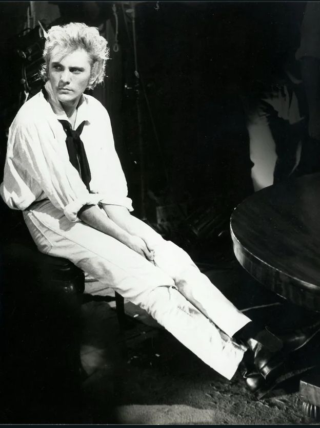 A man with blond hair wearing sailor’s cloths sits with his ankles crossed. 