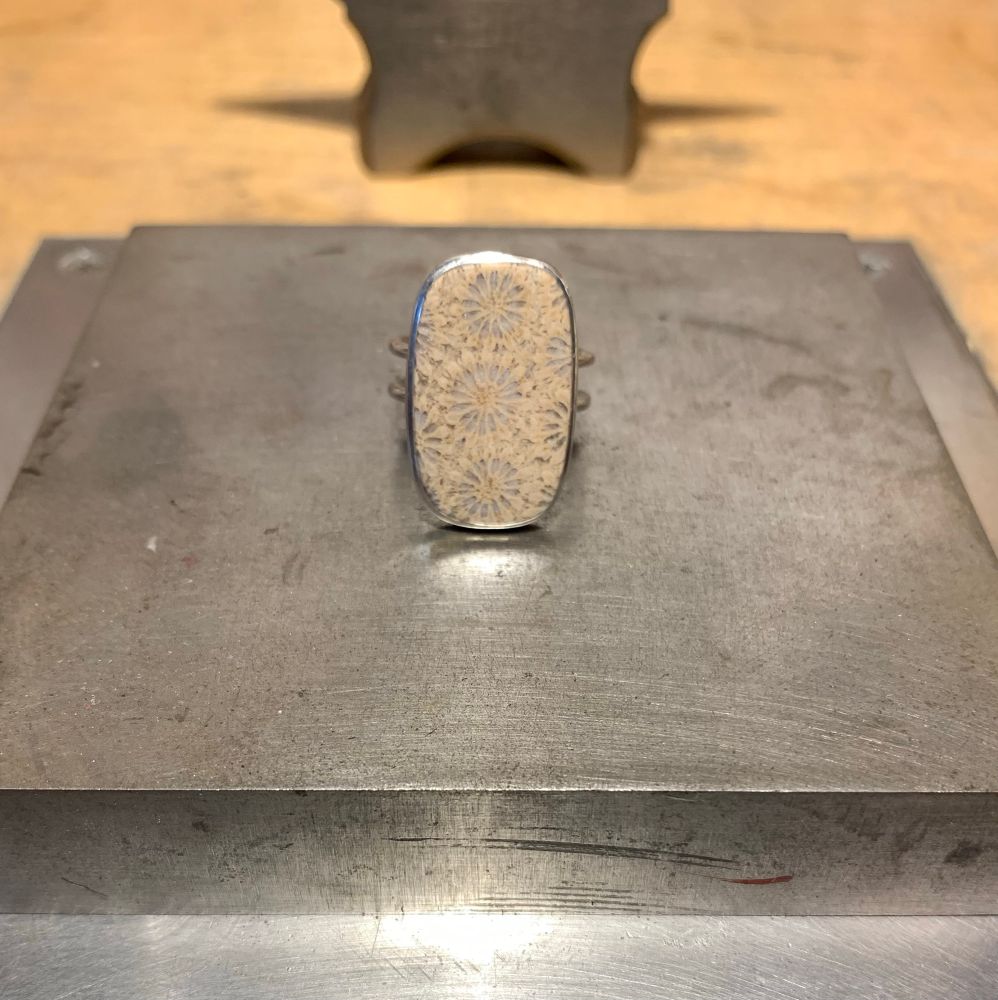 A coral fossil cabochon with pastel colors of lavender and peach set in a sterling silver ring, sitting on a jewelers bench block.