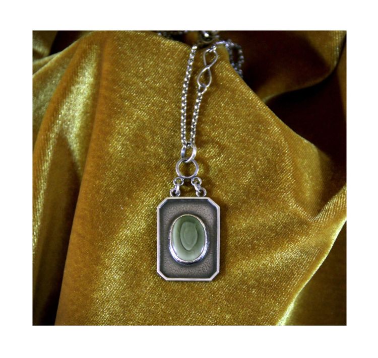 A green striated Jasper is set in an octagonally framed pendant suspended by two chains from a central ring. It lays against a gold velvet backdrop. 
