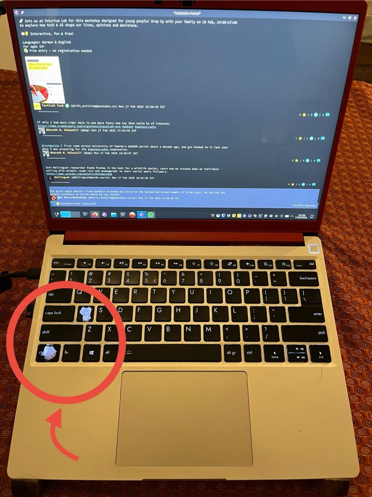 An open Framework laptop displaying a terminal window with text and email excerpts, along with a blurred workshop flyer on the screen. The keyboard shows worn keys, particularly the 'a' and 'ctrl' keys. The laptop is on a patterned surface.