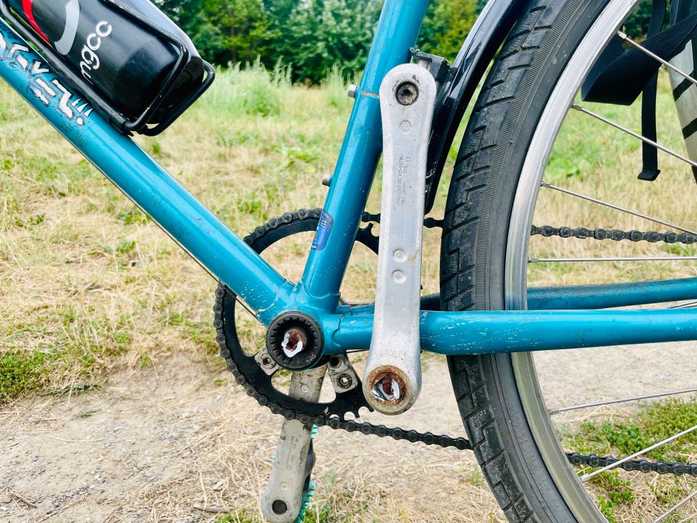 A closeup of the left side of the bottom bracket of a blue 90s mountain bike. The spindle has completely sheared. I’ve propped the left crank, which contains the rest of the spindle, on the frame. 