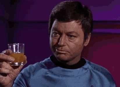 Dr. McCoy from Star Trek raises a glass of an orangish liquid.  He nods approvingly.  He has a smirk on his face.