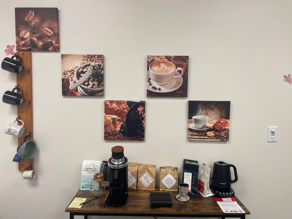 Bottom center of the photo contains a wood top table with a coffee grinder, digital scale, small cup made to look like birch bark, an electric kettle, a chemex pour over vessel, and 8 varieties of coffee beans sitting on top of it. On the wall center frame is 5 different square canvas prints of coffee accoutrements (coffee beans pouring down, two coffee grinders with beans in and around, and two different coffee cups, one with pastries haphazardly placed nearby ) arranged in an asymmetric up and down pattern. On the wall to the left is a mug rack containing 2 black mugs, one with a dragoon with the colors of the Pan flag, a large green and black mug, and 2 small espresso mugs.
