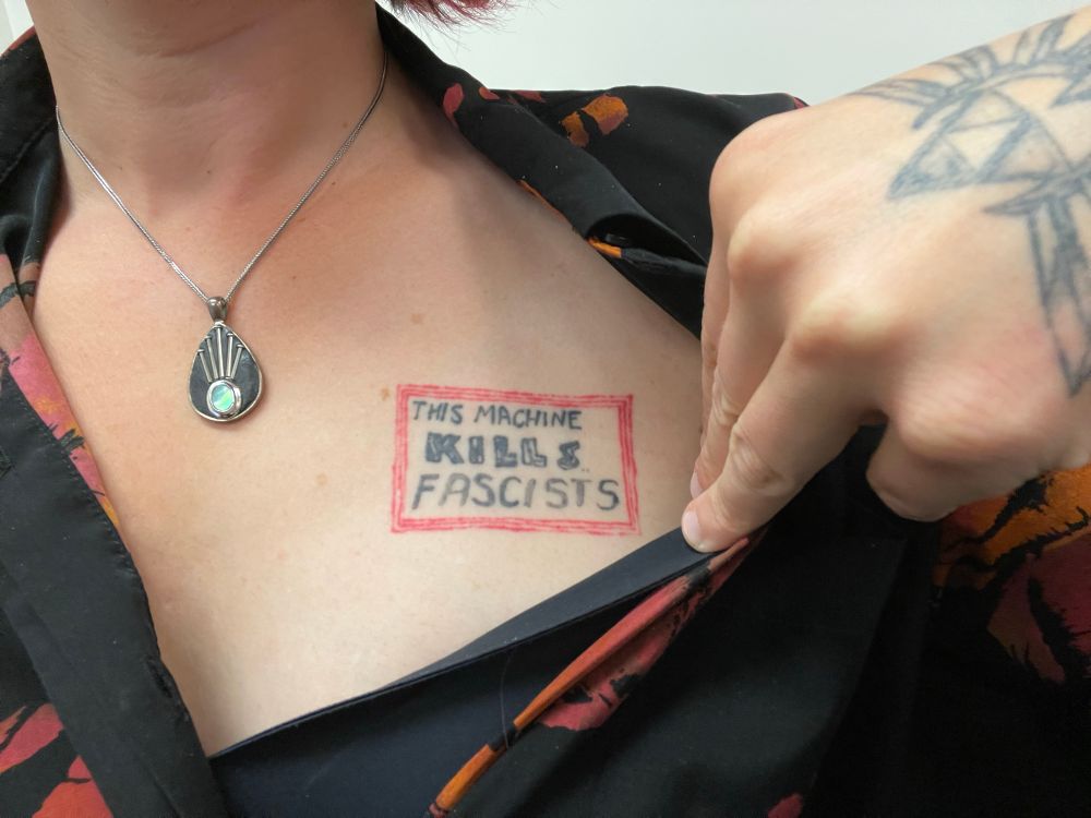 Close up of a white woman with a “This machine kills fascists” tattoo being shown. The tattoo is in the style of Woody Guthrie’s famous stickers that he put on his guitars.