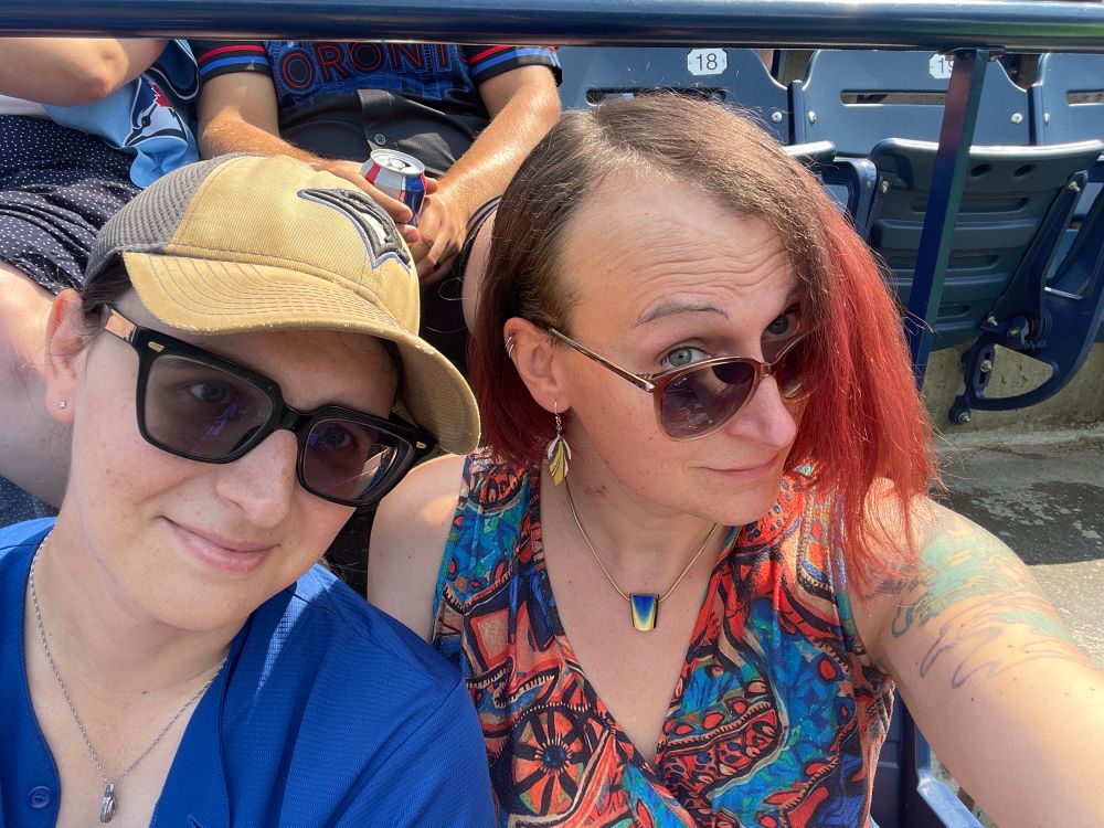 Two women wearing sunglasses looking at the camera. Woman on the left is wearing Blue Jays fan Paraphernalia, woman on the right (me) is wearing a colorful dress with an abstract pattern. Photo was taken at a baseball stadium, and bleacher seats are seen behind them.