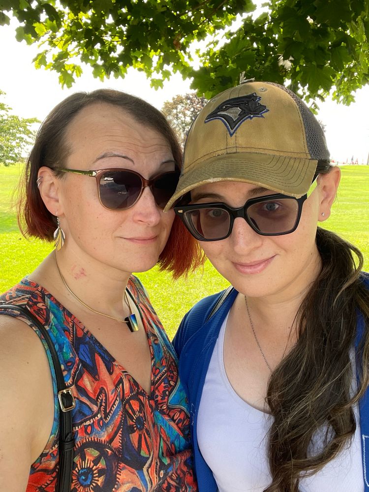 Two women wearing sunglasses looking at the camera. Woman on the right is wearing Blue Jays fan Paraphernalia, woman on the left (me) is wearing a colorful dress with an abstract pattern. Behind them is a grassy field and a lake, with leafy tree branches at the top of the photo.