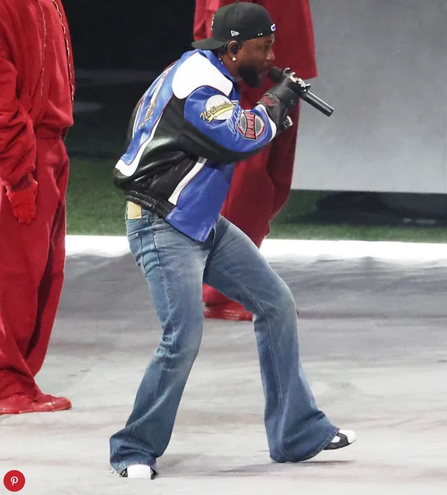 Rapper Kendrick Lamar, in a pair of throwback boot cup jeans, and white tipped tennis shoes. He sports a blue, black, and white leather jacket with Gloria on the front. He is also wearing a backwards, black baseball hat.