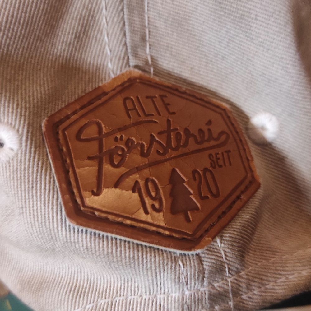 Das Bild zeigt ein detailreiches Nahaufnahme eines braunen Lederetiketts auf einer grauen Kappe. Das Etikett ist sechseckig und trägt die Aufschrift „Alte Försterei seit 1920" in einer stilisierten Schriftart. Ein kleines Tannenbaumsymbol ist ebenfalls abgebildet. Das Leder wirkt abgenutzt und leicht rissig, was einen vintage Look vermittelt.
