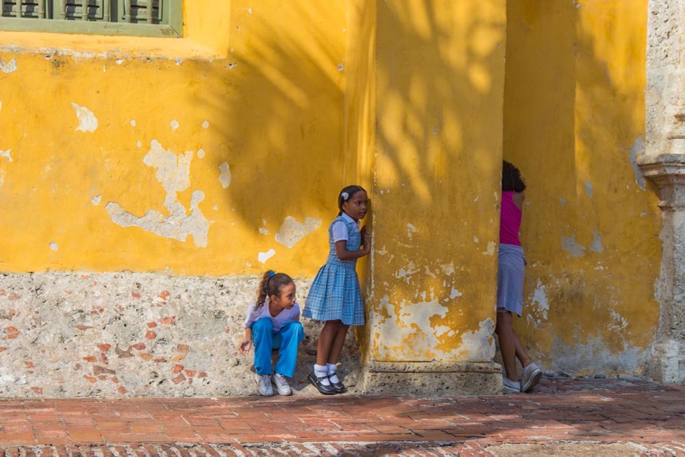 Dos niñas en uniforme de comegio se esconden tras una columna mientras otra cuenta en el juego de Escondidas. Es la pared amarilla de una iglesia caribeña
